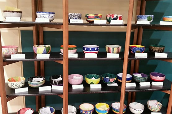 A collection of custom-painted empty bowls with paper descriptions in front of them line up on a shelf