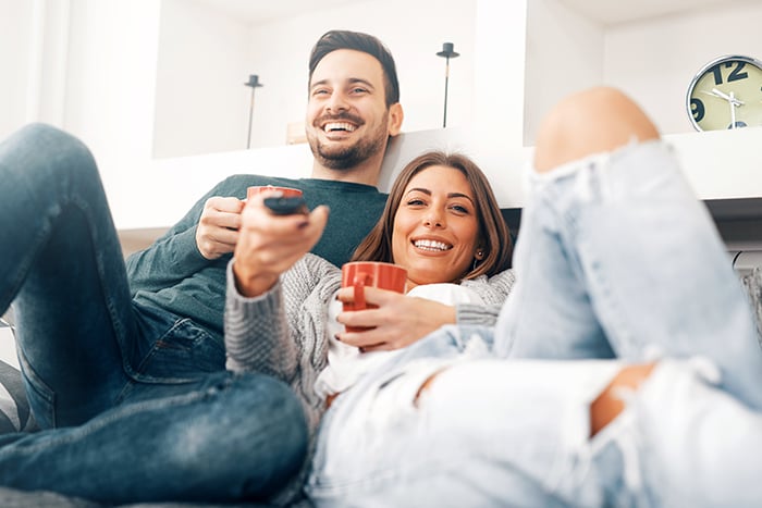 happy man and woman snuggle up on couch with coffee and watch tv