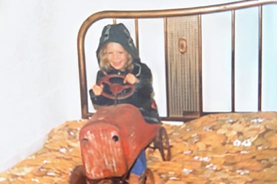 A young Kim Settel sits on her vintage red pedal car, which is parked on top of a bed