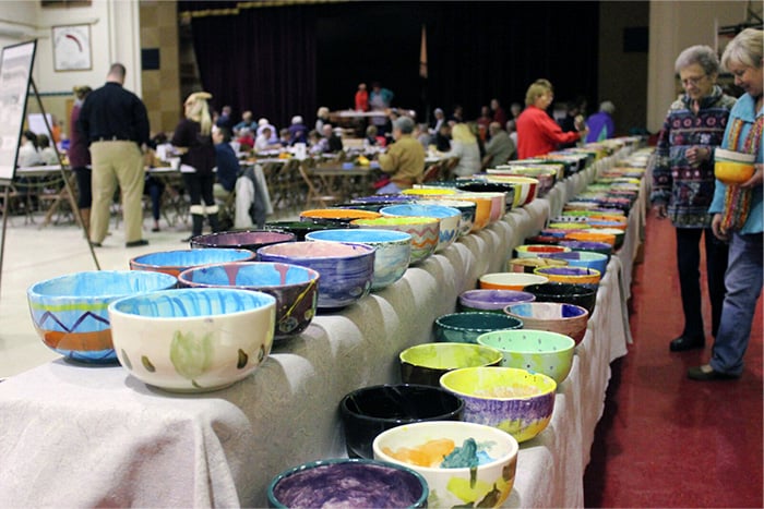 Dozens of custom-painted empty bowls line a long table as onlookers admire them from nearby