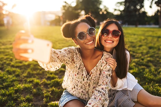 two girls sit in the sunshine outside while taking a selfie after reading a helpful cybersecurity article from gate city bank