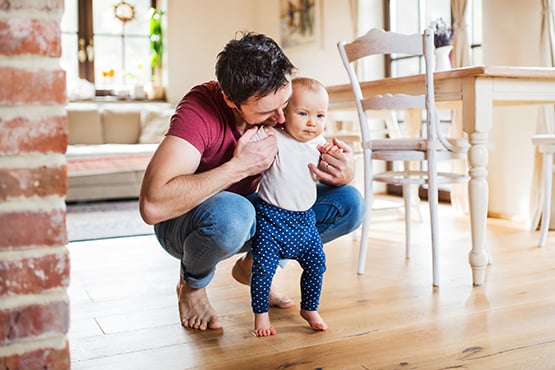 dad with messy hair kneels beside a blue-eyed toddler, holding her hands as she takes her first steps in their new home