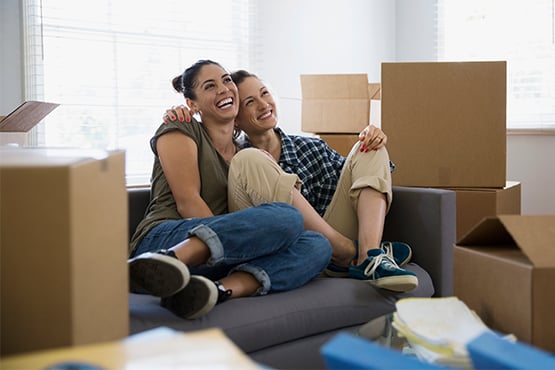 Two smiling women on couch surrounded by moving boxes who had wondered whether to rent or buy a new home with gate city bank