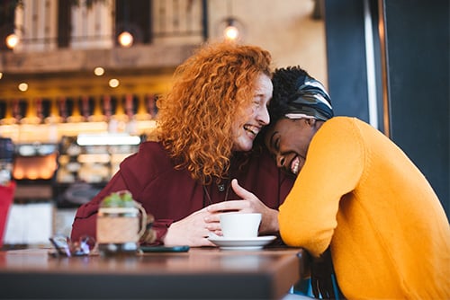 Two millennial women laughing over coffee, earning points at a local café thanks to Gate City Bank debit card rewards