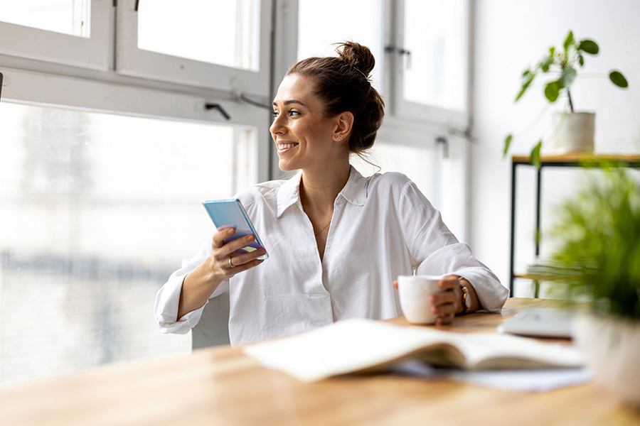 happy young woman sits at desk and looks out window while holding phone