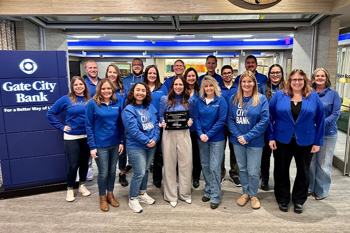 a large group of blue-shirted Gate City Bank mortgage team members huddle around the NDHFA Affordable housing Award for 2023