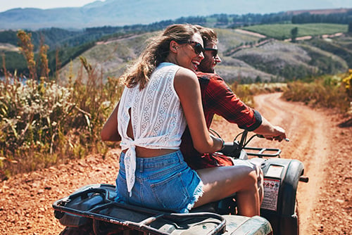 Laughing couple wearing sunglasses and riding a quad in the summertime, dusting up gravel on western ND trails