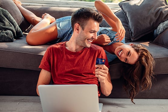 Laughing couple on a couch with their laptop, comparing the perks of a Gate City Bank debit card vs credit card