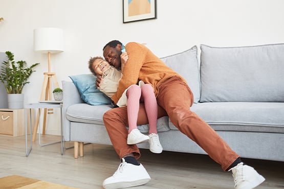 father and child laughing on sofa in living room after protecting home with additional homeowner’s insurance