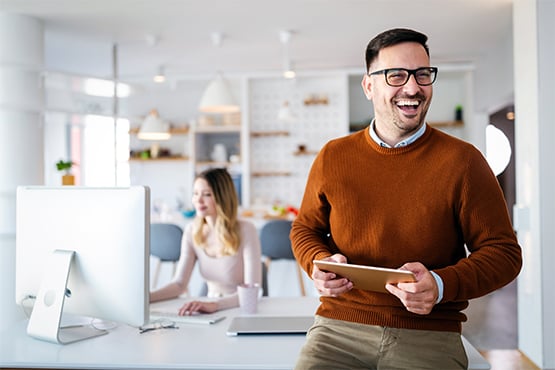 joyful male business owner uses tablet to gain easy online access to his business credit card while female employee works in the background