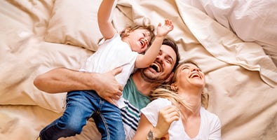 mother father and young child laugh as they fall back after a fun pillow fight