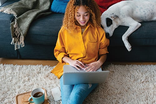 Young woman with curly hair, accessing her Preferred Perks Checking benefits on a laptop while sitting on living room floor
