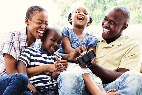 a young family of four laughs on the couch as they look at a phone