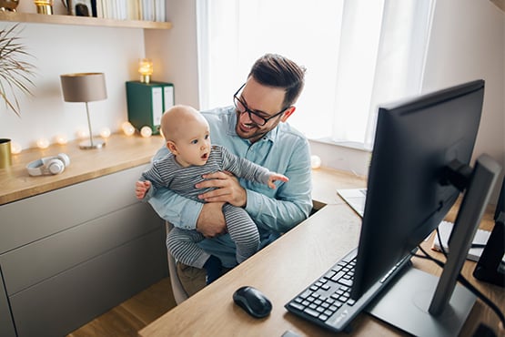smiling father holds toddler son at kitchen table and reads Gate City Bank’s article on reasons for a mortgage refinance while mom washes dishes