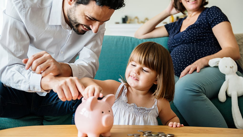little girl places coins into a piggy bank to save for future with happy mom and dad looking on