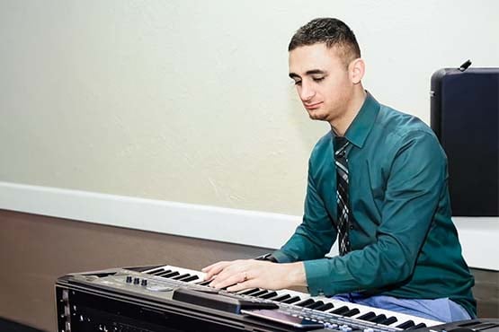 Wearing a teal dress shirt and tie, Lezan Tahir sits down to play an electronic keyboard