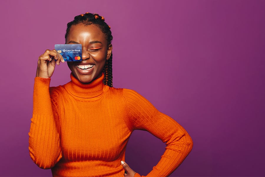 Close-up of a young woman in an orange turtleneck, smiling and holding her Gate City Bank debit card over one eye