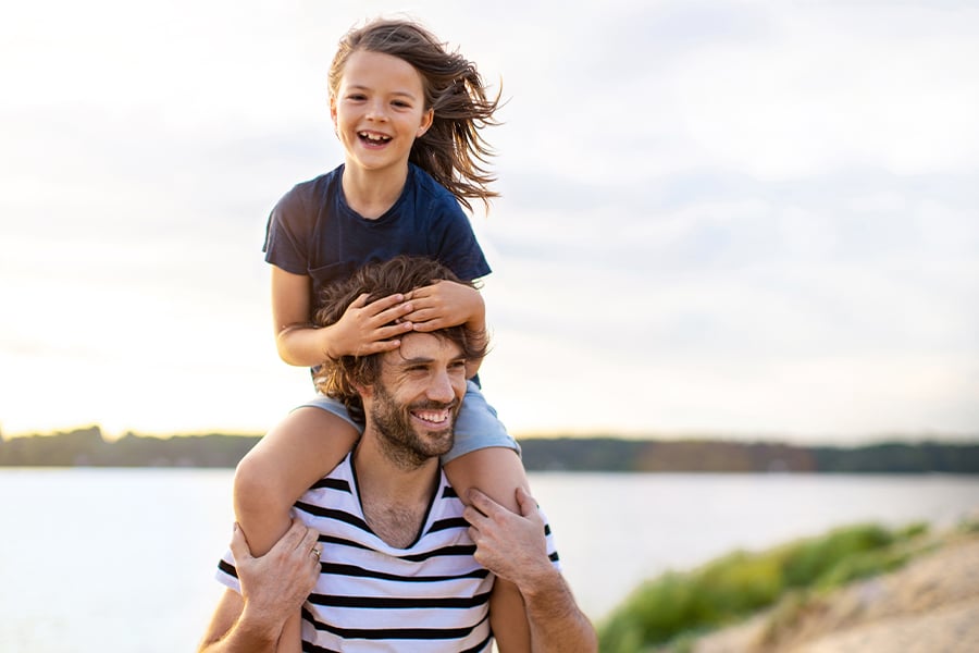 Laughing girl sitting on her father’s shoulders, walking along the shore at their lake home in Minnesota