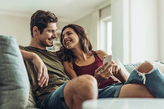 happy young couple sits on couch and smiles while woman holds phone