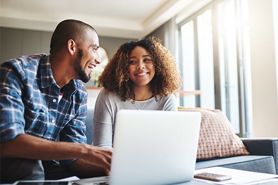 Young couple on sofa with laptop, trying Gate City Bank’s mortgage calculator to estimate their new house payment in Fargo, ND