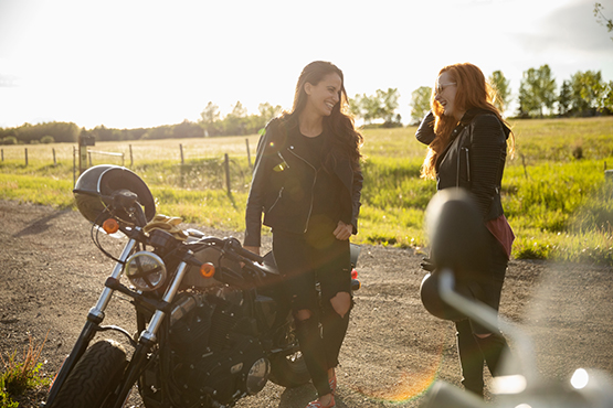 two girls laughing beside their motorcycles on a country road