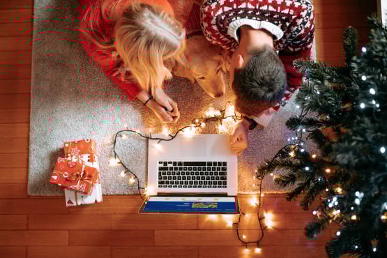 A couple works together to plan their holiday budget under the light of a Christmas tree, while their dog lounges between them