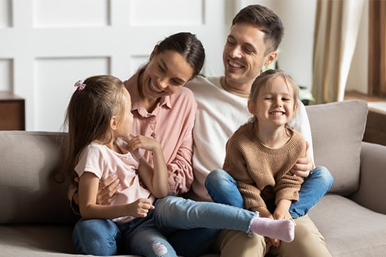 happy young parents and daughters sit on living room couch after checking out Gate City Bank's tips to set a household budget