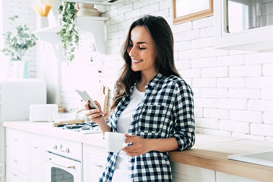 woman in kitchen holds coffee and uses phone to enjoy a low minimum account balance