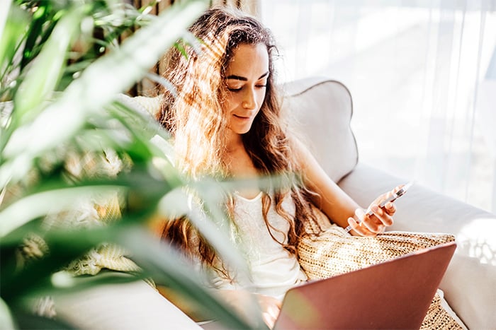 happy sun lit young woman sits on chair with computer and uses phone to order checks