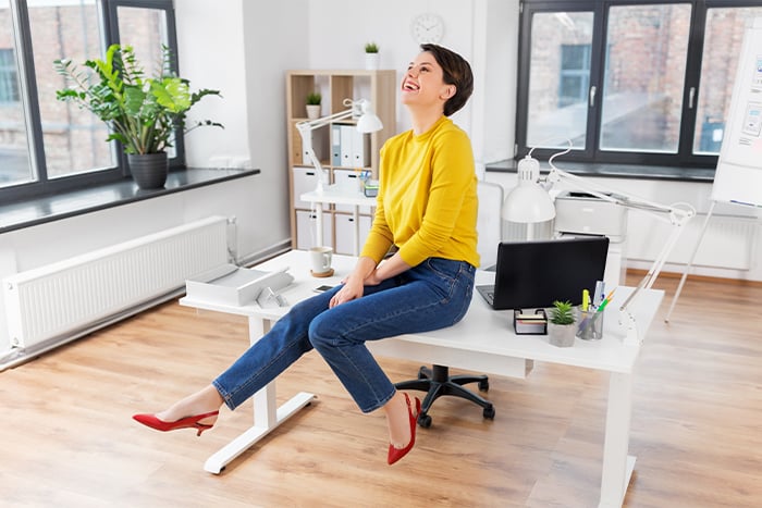 happy woman business owner sits on edge of desk in brightly lit office after enjoying remote deposit capture services from Gate City Bank