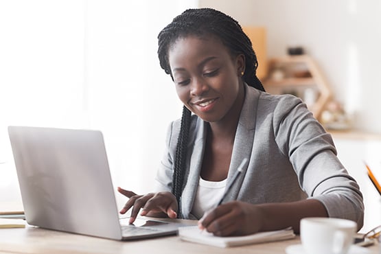 smiling woman works on her laptop and uses notepad and pencil to achieve financial success with gate city bank