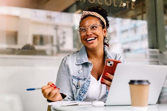 A happy woman sits with her devices at a coffee shop after learning about paying off student loan debt from Gate City Bank