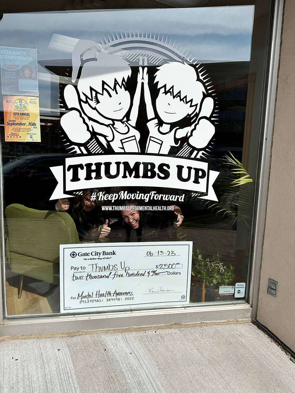 two excited women with nonprofit Thumbs Up in Elk River, MN showcase Gate City Bank’s big check at their front window