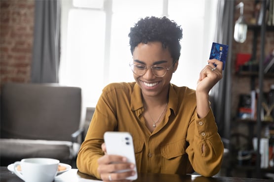 Cheerful millennial holding her Gate City Bank debit card in one hand while online shopping on her phone