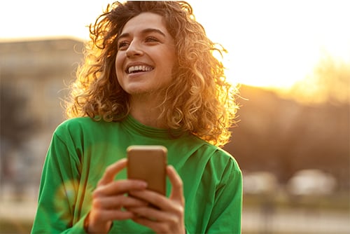 happy young woman in green shirt holds phone outside after reading FAQs on scammers