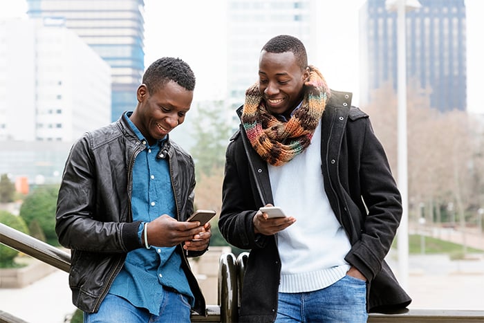 two happy male friends hold phones within a city and send money online with Zelle