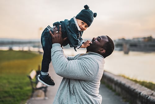 Toddler boy in a winter hat, smiling while being lifted into the air by his dad along the riverbank in Elk River, MN