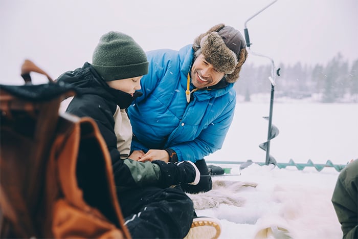 dad in fuzzy bomber hat smiling at his son on a frozen Minnesota lake, excited to talk about ice house loans with Gate City Bank