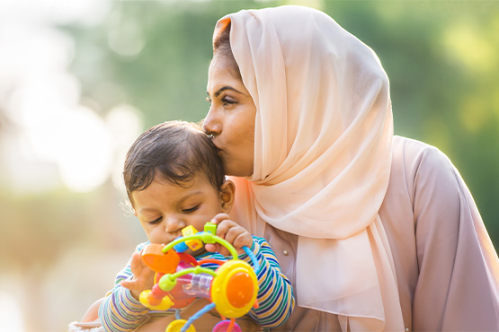Beautiful mother new to America, kissing her toddler son’s hair while sitting together at Gooseberry Park in Moorhead, MN