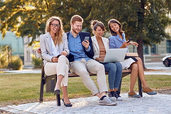 four happy young professionals sit on park bench with devices and enjoy online banking