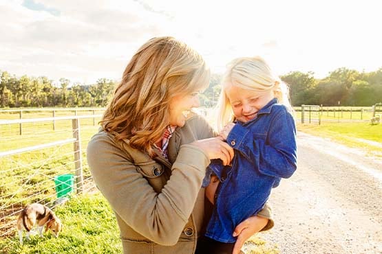 Smiling mom holds her young blonde daughter on her hip, standing on a gravel road on their farm outside Mayville, ND