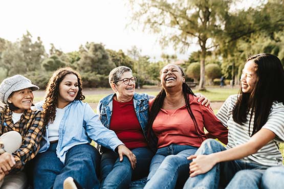 Five laughing women of different ages, sharing stories while sitting on the grass in jeans in Roosevelt Park in Minot, ND