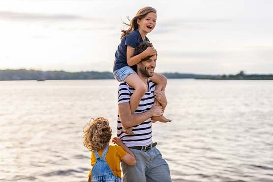 Young brunette girl laughing on her dad’s shoulders as they walk along the edge of the water in Devils Lake, ND