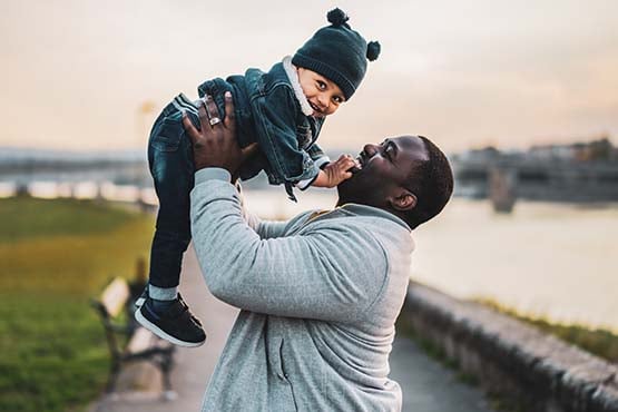 Toddler boy in a winter hat, smiling while being lifted into the air by his dad along the riverbank in Elk River, MN