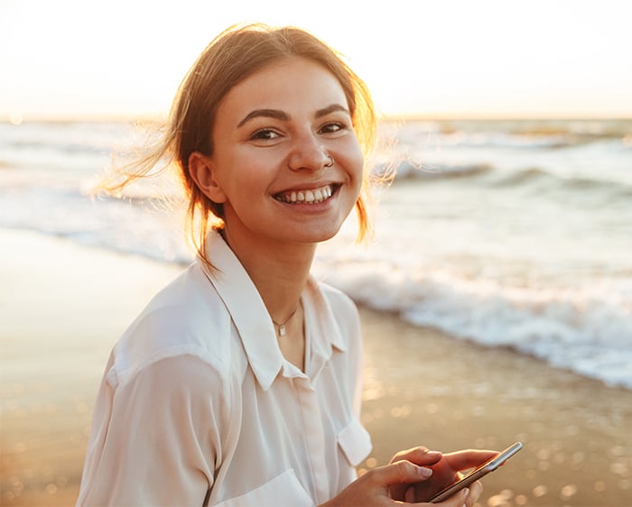 happy young woman enjoys mobile banking on her smartphone while she strolls along an ocean beach with a sunset in the backdrop